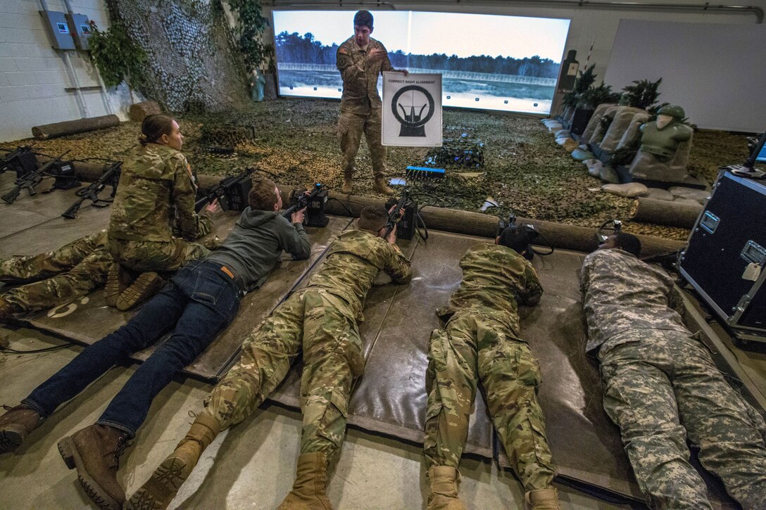 Cadets lie prone with rifles while looking up at a soldier holding a diagram in a room designed to look like an outdoor range.