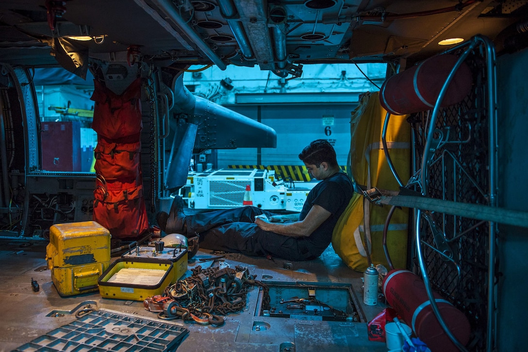 A sailor sits and reads a list on the floor of a ship's hangar bay, illuminated in blue.
