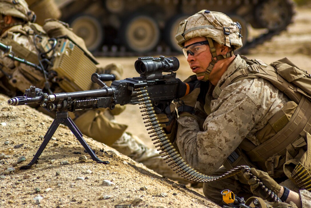 A Marine aims a machine gun while lying behind a berm in desert terrain.