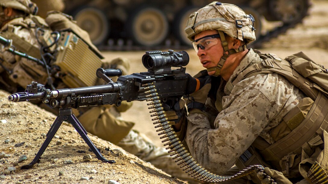 A Marine aims a machine gun while lying behind a berm in desert terrain.