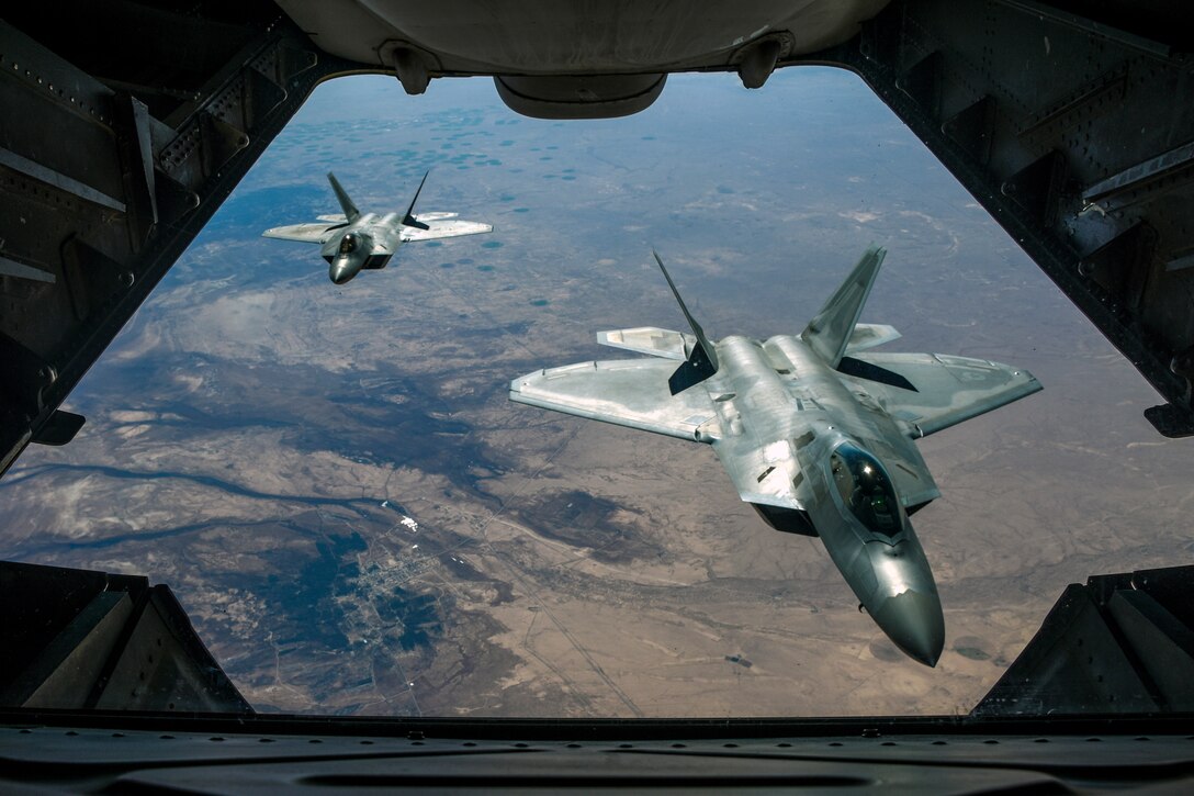 Two fighter jets, viewed from an opening in a third aircraft in front of them, fly over brownish terrain.