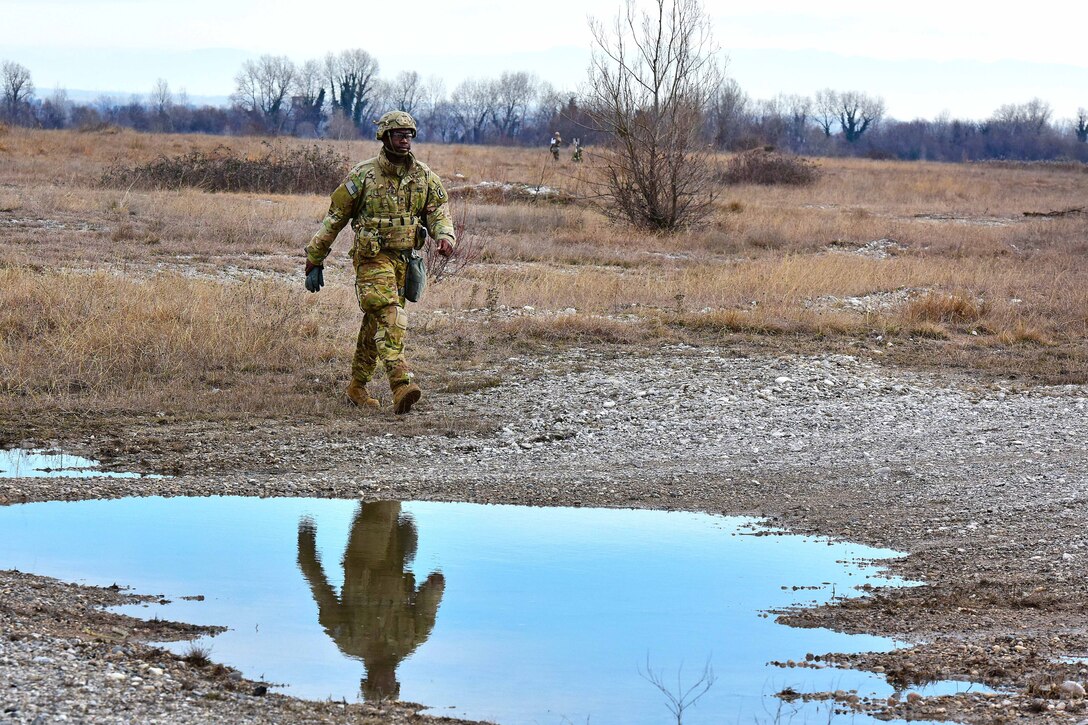 A soldier's reflection is shown in a puddle as he walks on rocky terrain.