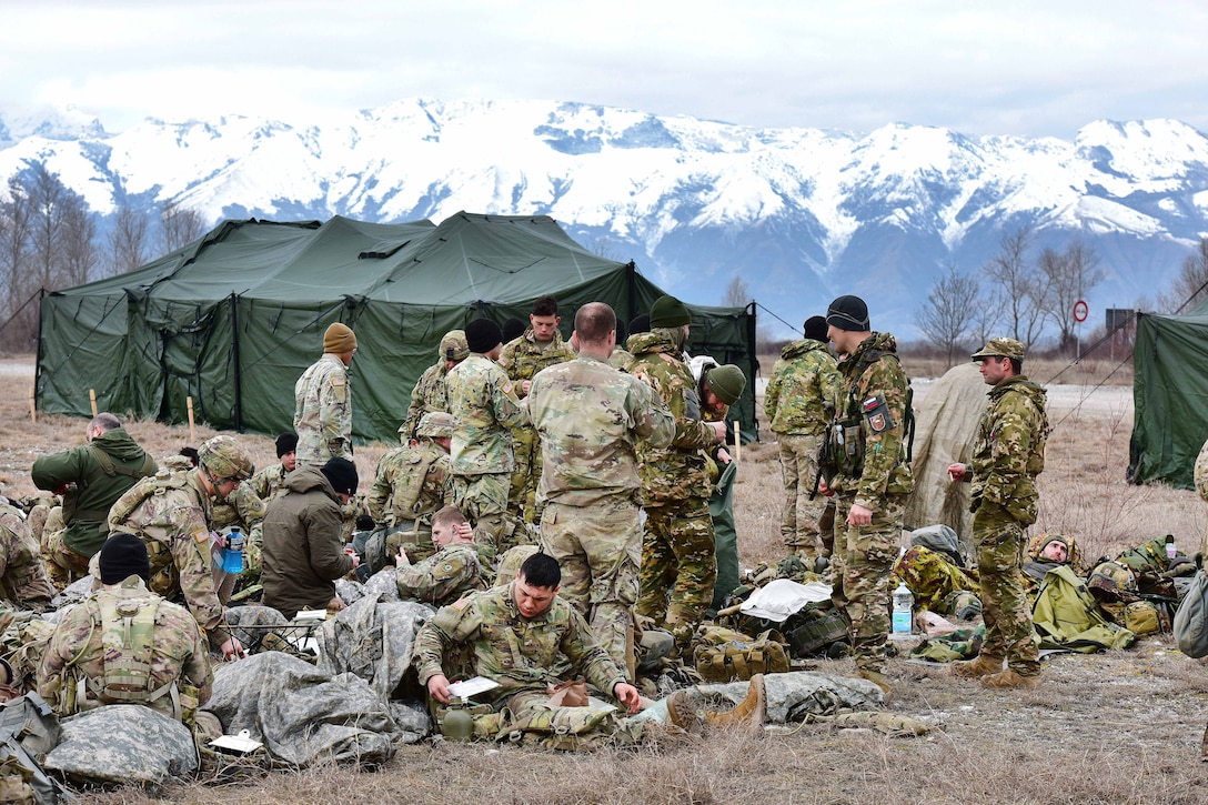 U.S., Italian and Slovenian paratroopers sit and stand outside near a tent.