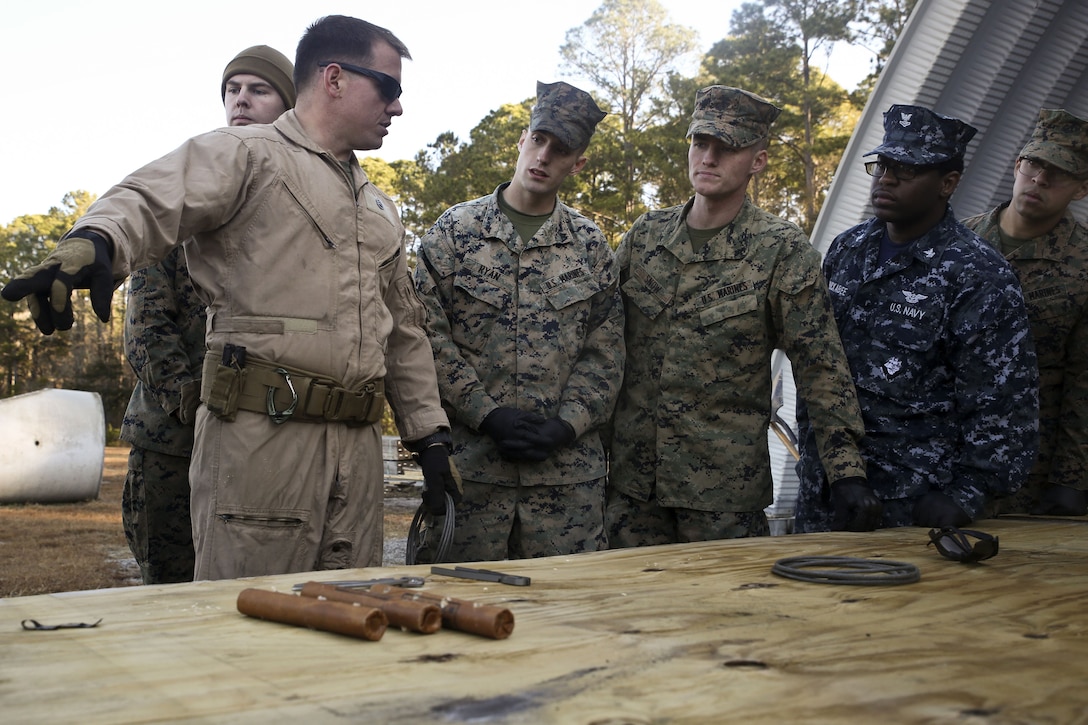 A Marine Corps instructor speaks to Marines and sailors in front of a table.