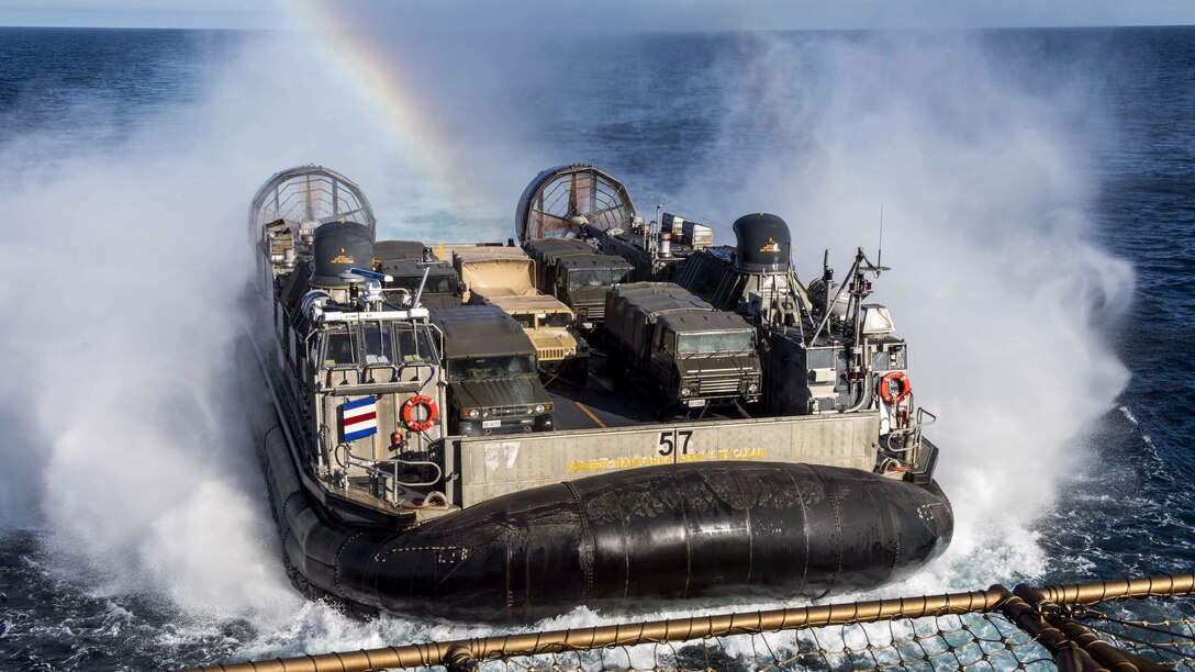 An air-cushioned landing craft lapproaches a ship as a rainbow forms over it.