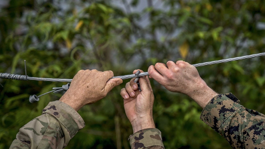 The hands of a soldier and a Marine fix a wire on a fence.