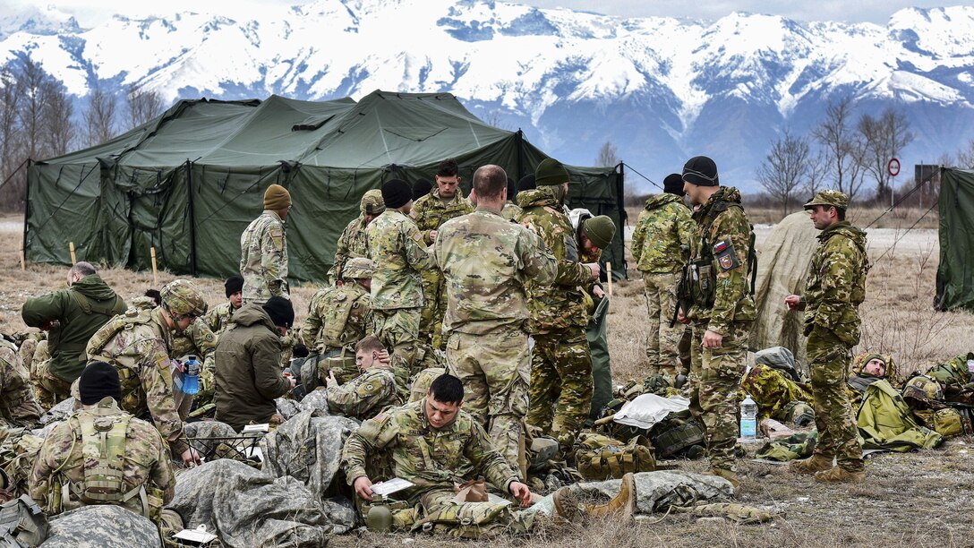 U.S., Italian and Slovenian soldiers relax outside with mountains behind them.