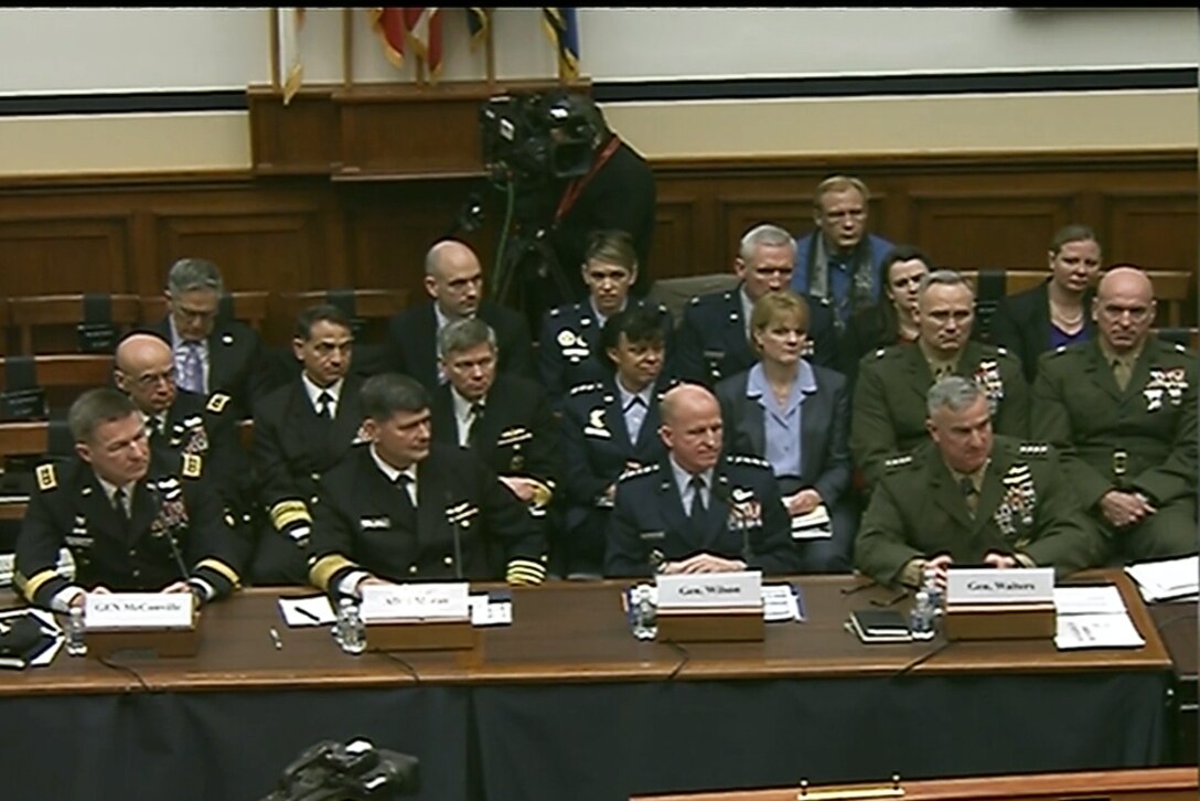 Military officials sit behind a desk with other civilians and military sitting behind them.