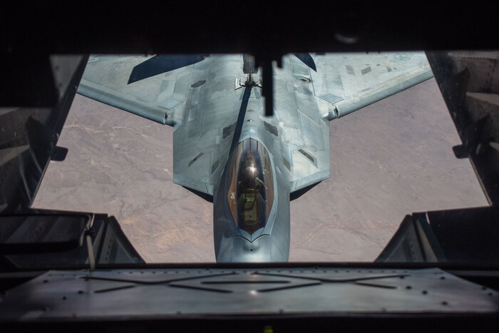 A fighter jet, viewed from an opening in a second aircraft above it, flys over brownish terrain.