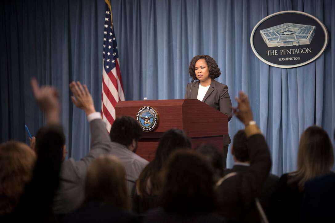 A person speaks from behind a podium with people in the crowd raising their hands.