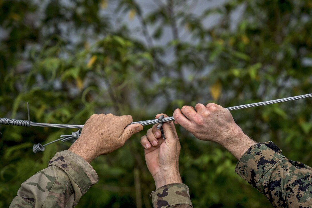 The hands of a soldier and a Marine fix a wire on a fence.