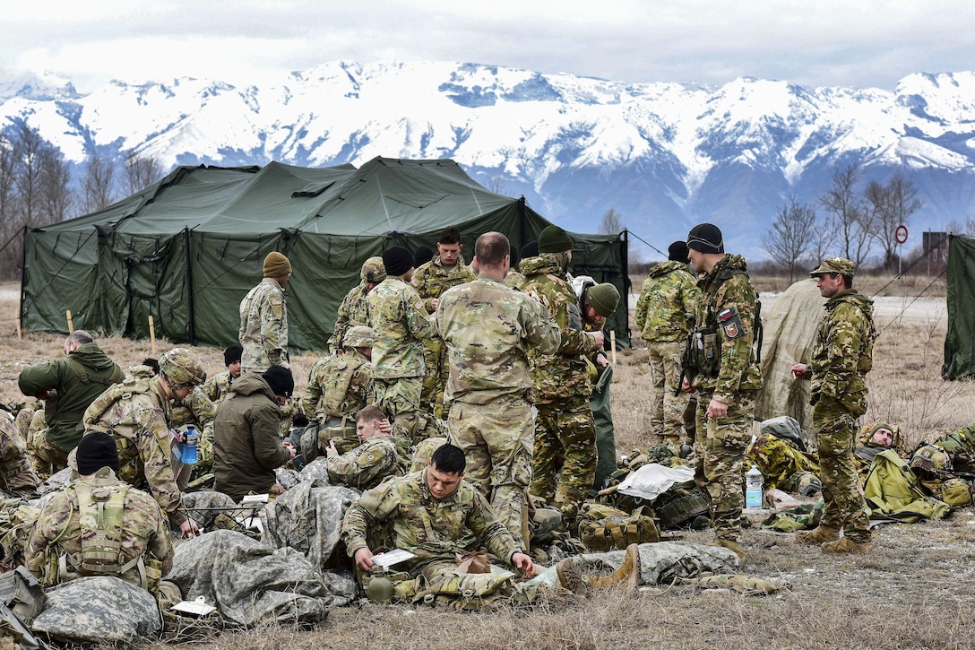 U.S., Italian and Slovenian soldiers relax outside with mountains behind them.