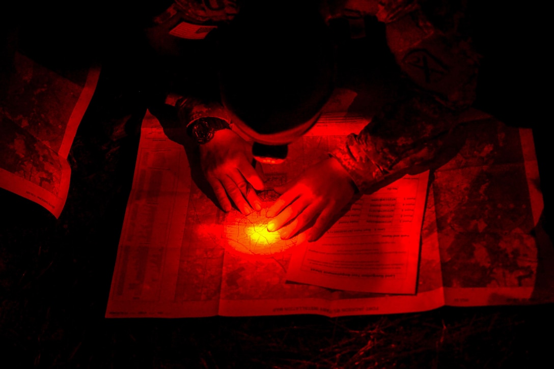 A soldier uses a headlight while crouching over a map at night, illuminating the scene in red.