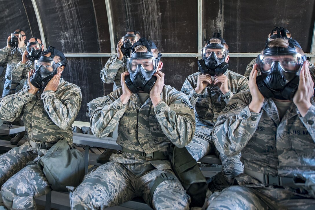 Airmen seated on metal bleachers place their hands over side vents on gas masks they are wearing.