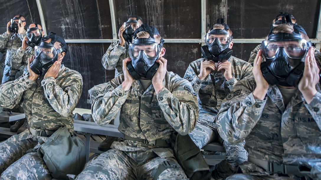 Airmen seated on metal bleachers place their hands over side vents on gas masks they are wearing.