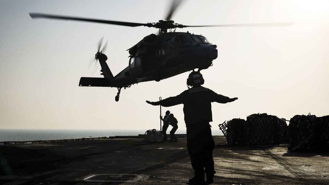 A sailor, shown in silhouette, signals to a helicopter as two others manipulate the aircraft's cargo line.