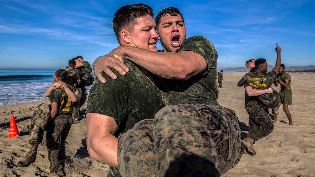 One Marine carries another on a beach, as other pairs of Marines do the same in the background.