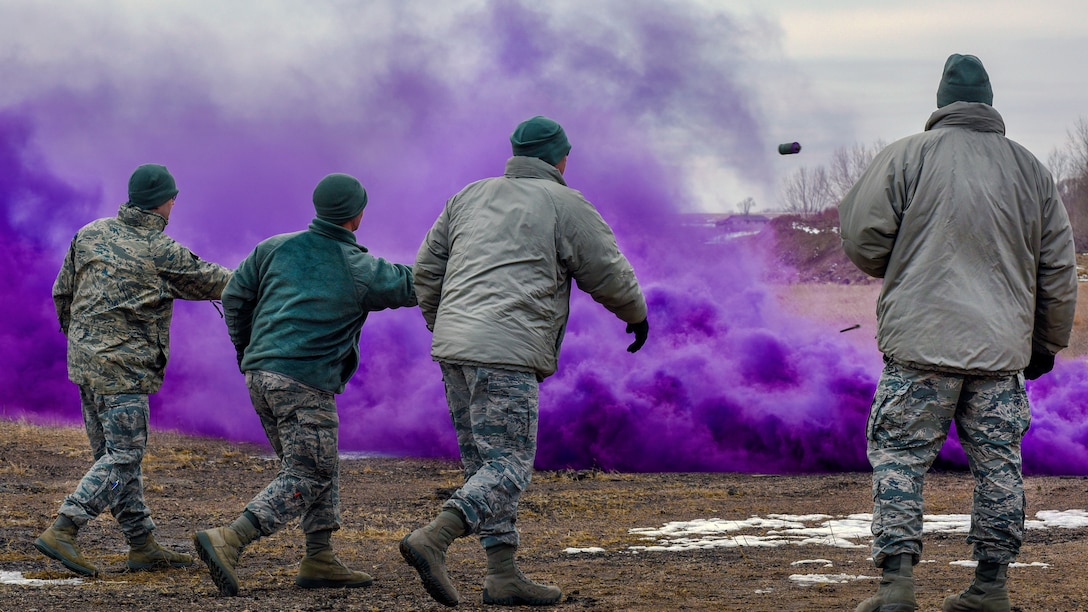 Three airmen in a row throw projectiles as a cloud of purple smoke rises in front of them.