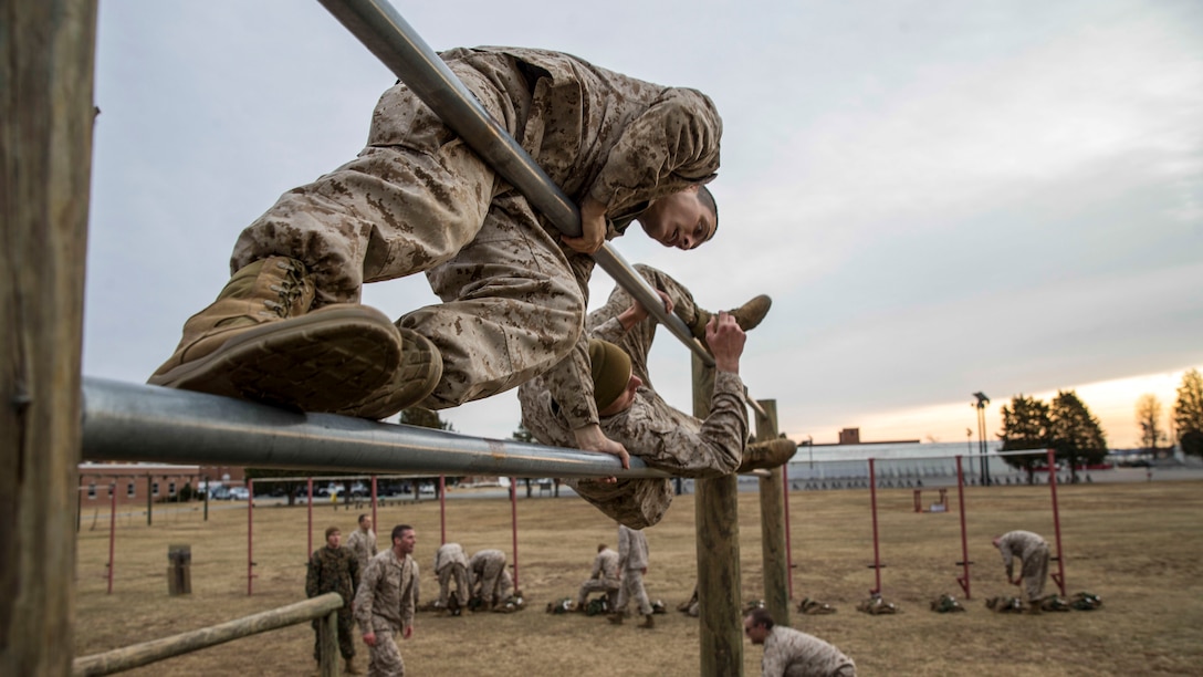 A Marine leans to one side while scaling a metal bar obstacle alongside another Marine at an outdoor course.