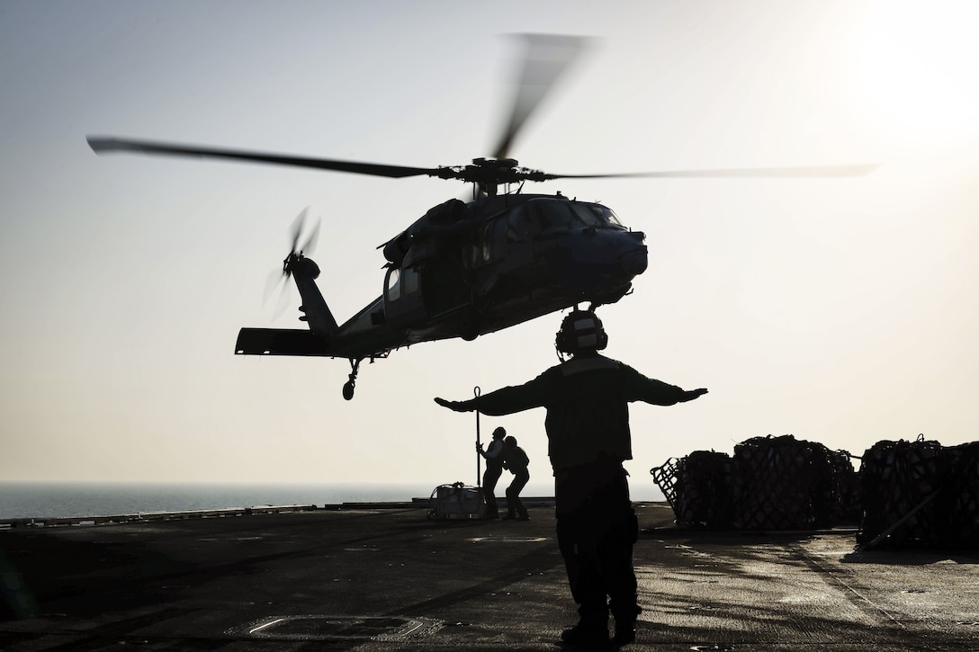 A sailor, shown in silhouette, signals to a helicopter as two others manipulate the aircraft's cargo line.