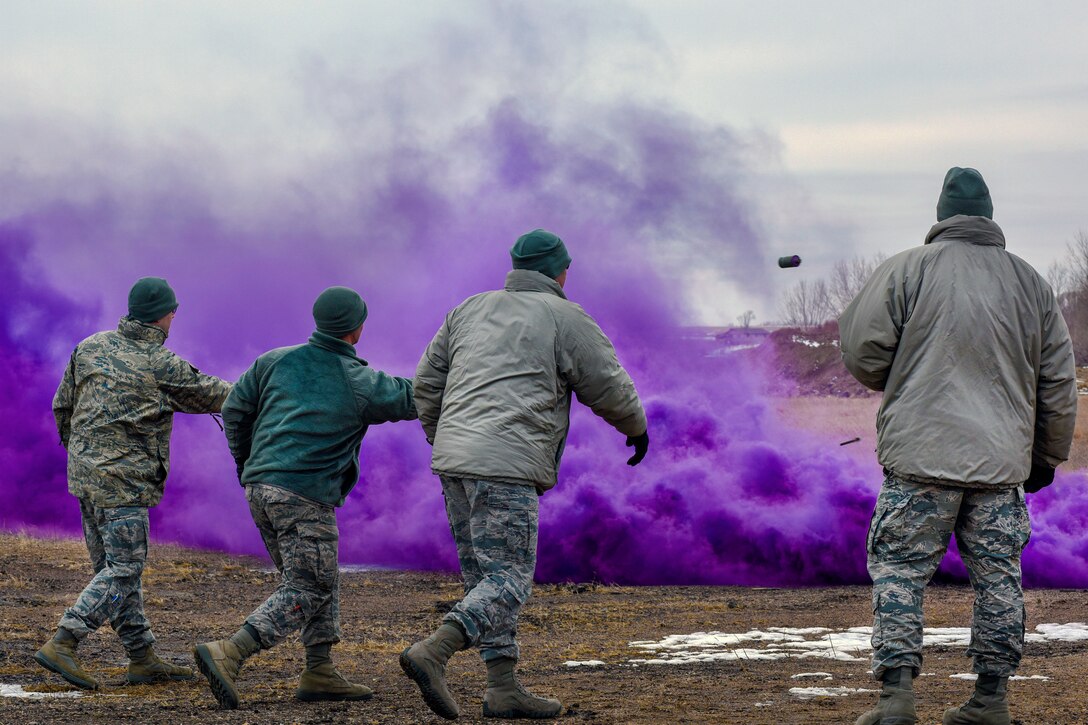 Three airmen in a row throw projectiles as a cloud of purple smoke rises in front of them.