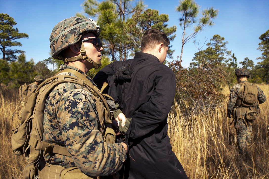 A Marine holds a detainee's arms behind their back as they walk through tall grass.
