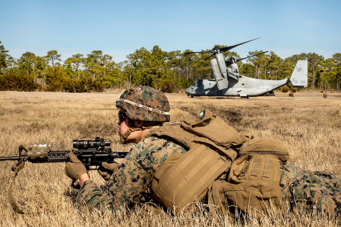 A Marine lays on the ground holding a rifle near an MV-22 Osprey.