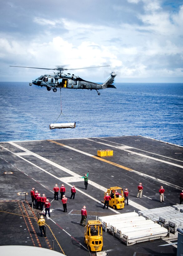 An MH-60S Seahawk helicopter carrying cargo hovers over a ship.