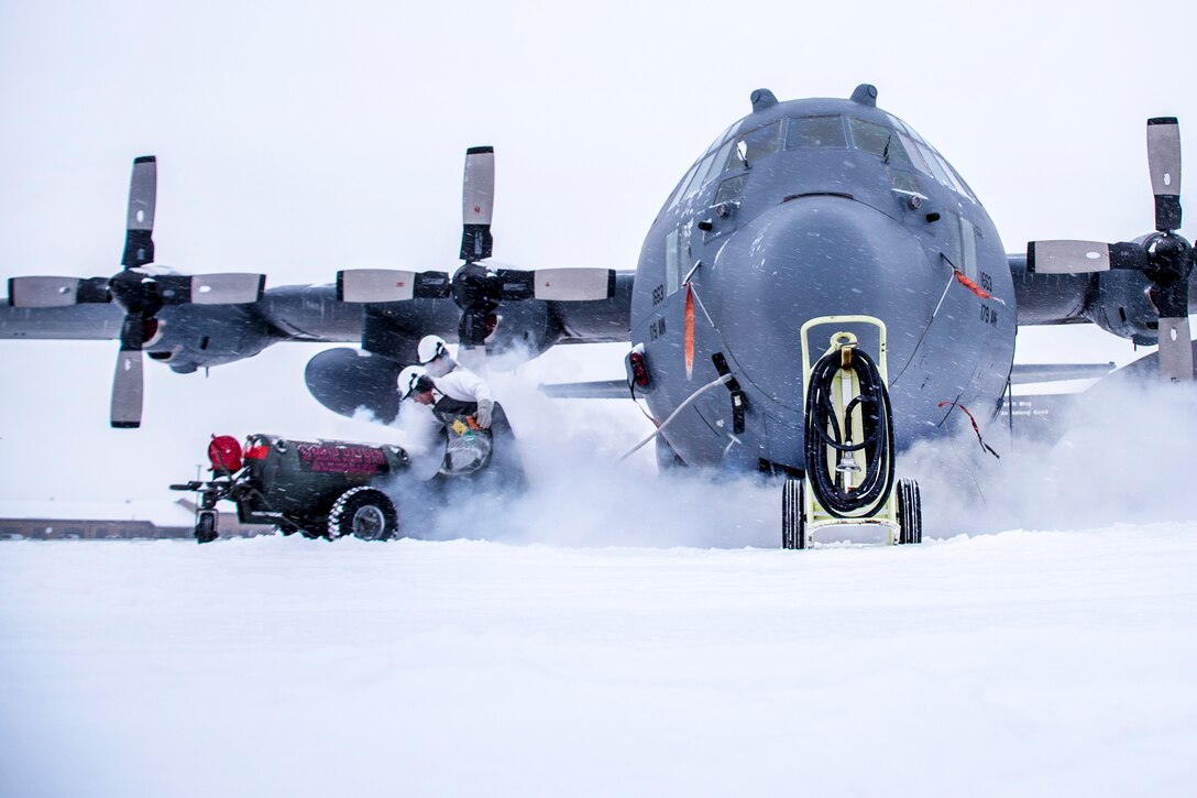 Airmen stand in blowing snow next to a Hercules aircraft.