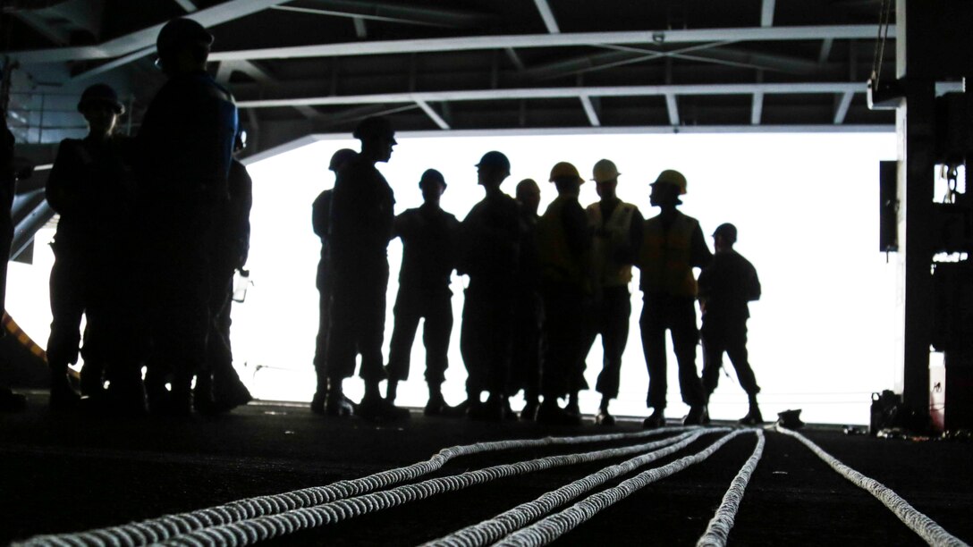 Sailors in silhouette stand in the hangar bay of a ship.
