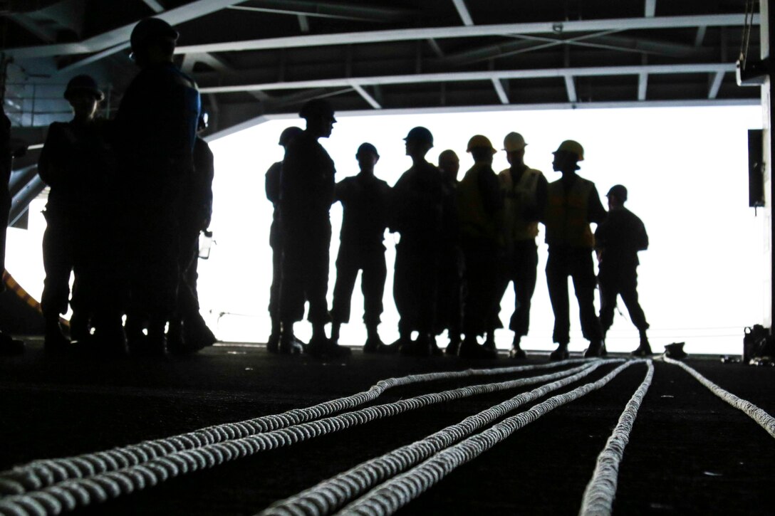 Sailors in silhouette stand in the hangar bay of a ship.