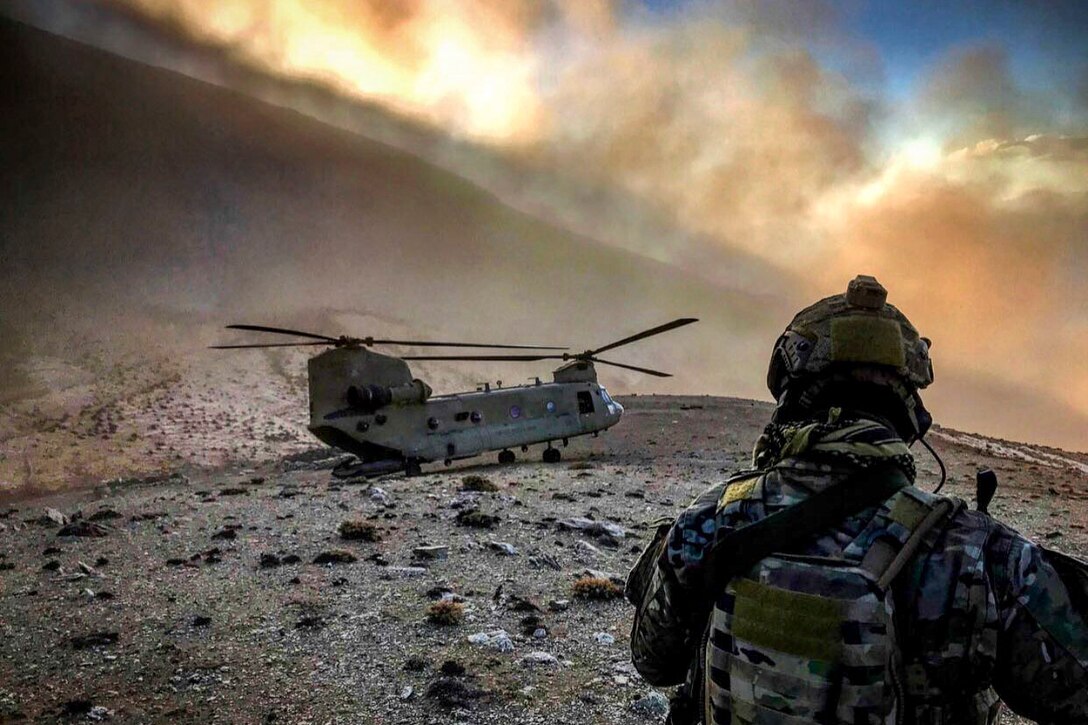 An airman, shown from behind, looks at a helicopter grounded on sparse, rocky terrain as clouds waft above.