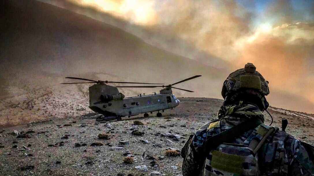 An airman, shown from behind, looks at a helicopter grounded on sparse, rocky terrain as clouds waft above.