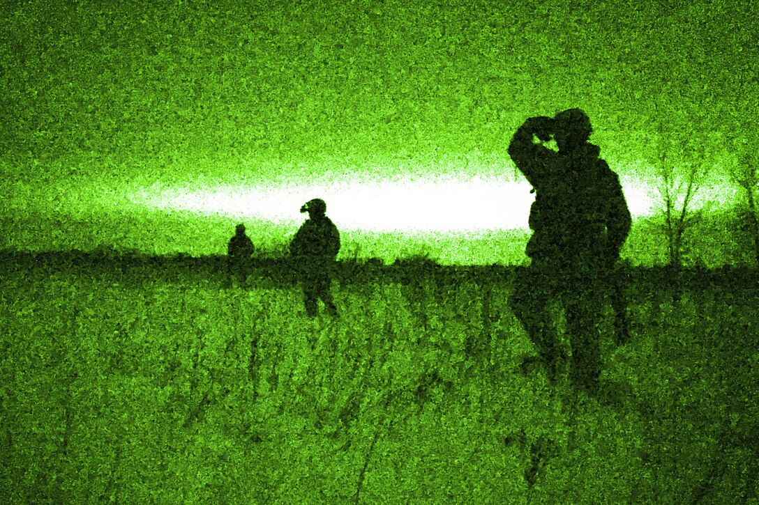 Airmen, shown in silhouette, walk in a field, illuminated in green from a night vision lens.