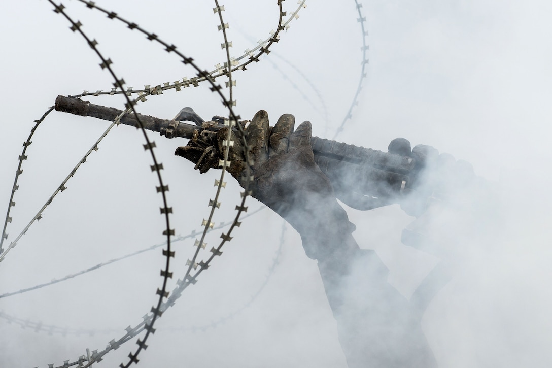 Hands holding a rifle hold up strings of barbed wire against a smoky backdrop.