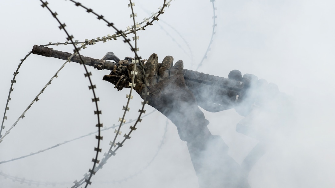 Hands holding a rifle hold up strings of barbed wire against a smoky backdrop