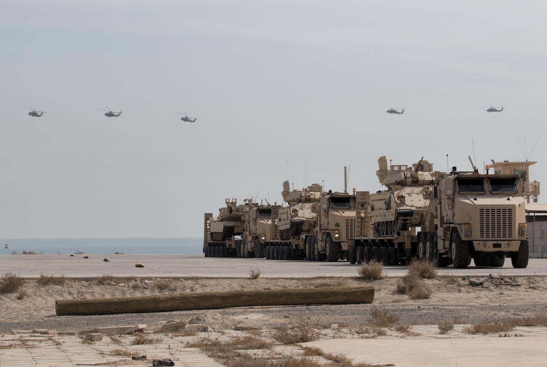 UH-60 Black Hawk helicopters fly over armored vehicles loaded on transport trucks during Army Day 2018, February 9, 2018, Kuwait Naval Base, Kuwait (U.S. Army/Ty McNeeley)