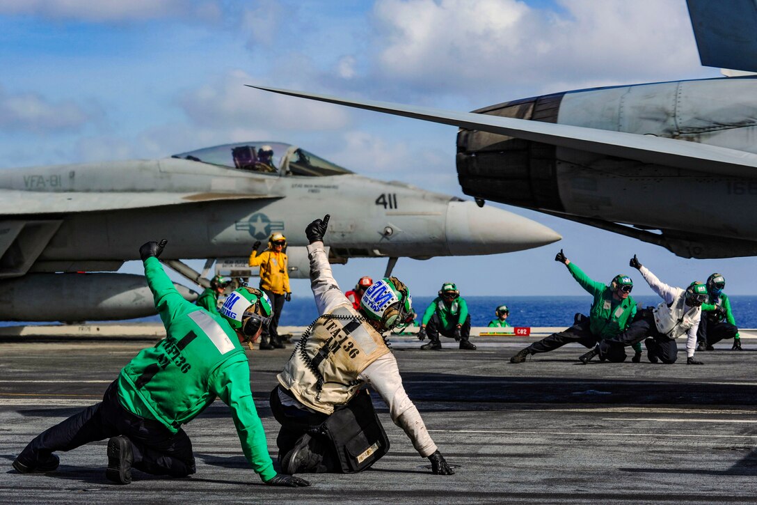 Two pairs of sailors kneel and give thumbs-ups on either side of an aircraft on a ship's flight deck.