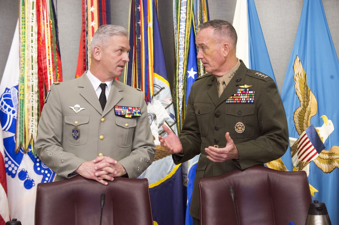 Marine Corps Gen. Joe Dunford stands and talks to his French counterpart in front of flags against a wall.