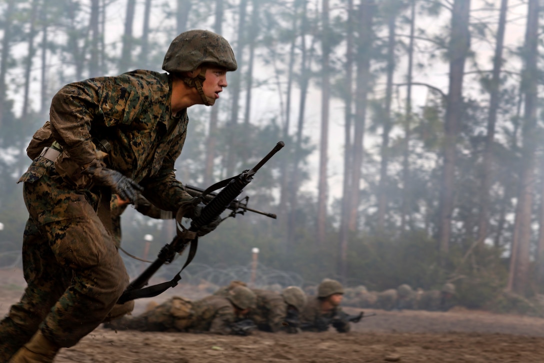 A Marine runs with a rifle in a wooded area.