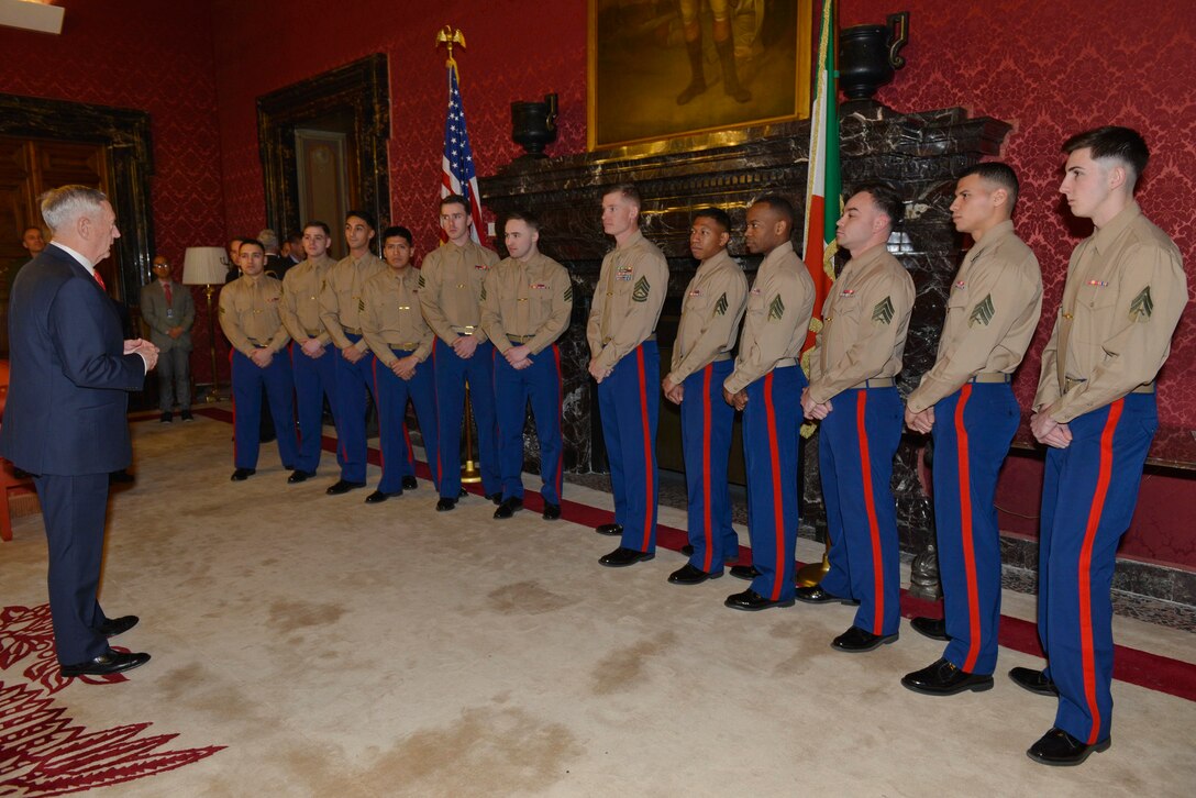 Defense Secretary James N. Mattis talks with 12 Marines standing in a row in a red-wallpapered room.