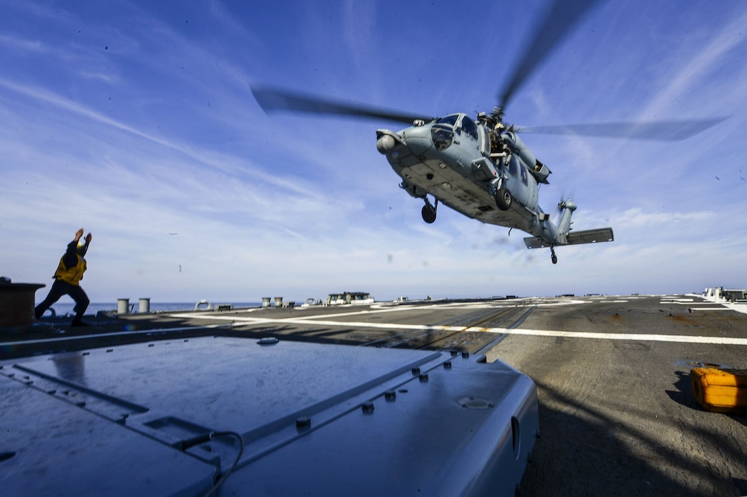 A sailor directs a helicopter to take off from a ship.