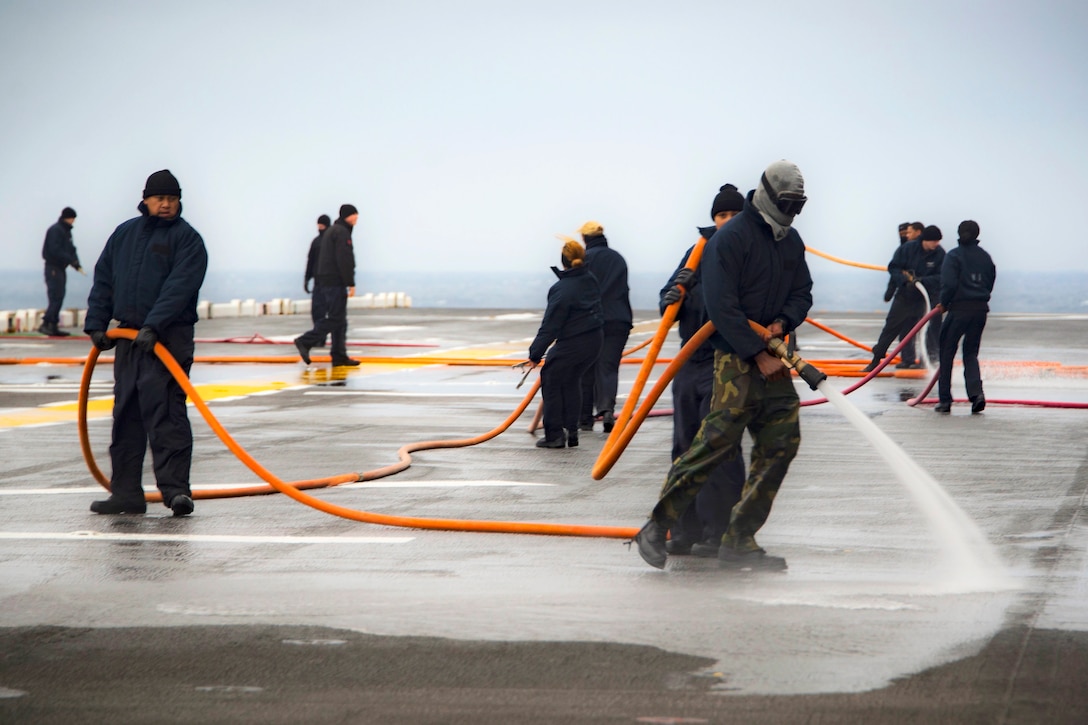 Sailors use long orange hoses to wash down a flight deck.