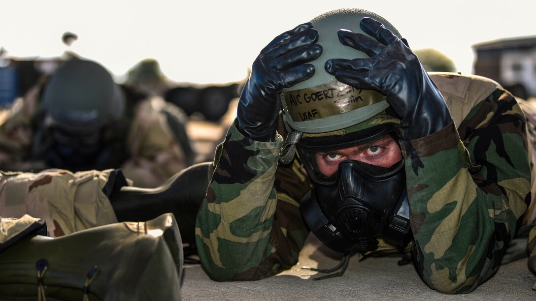 An airman in wearing a gas mask lies on the ground and places his hands on a helmet he's wearing.