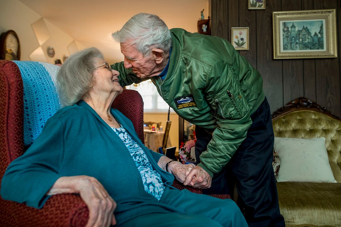 An elderly man leans in to kiss his wife, who holds her face up to his while sitting in a chair and holding his hand.