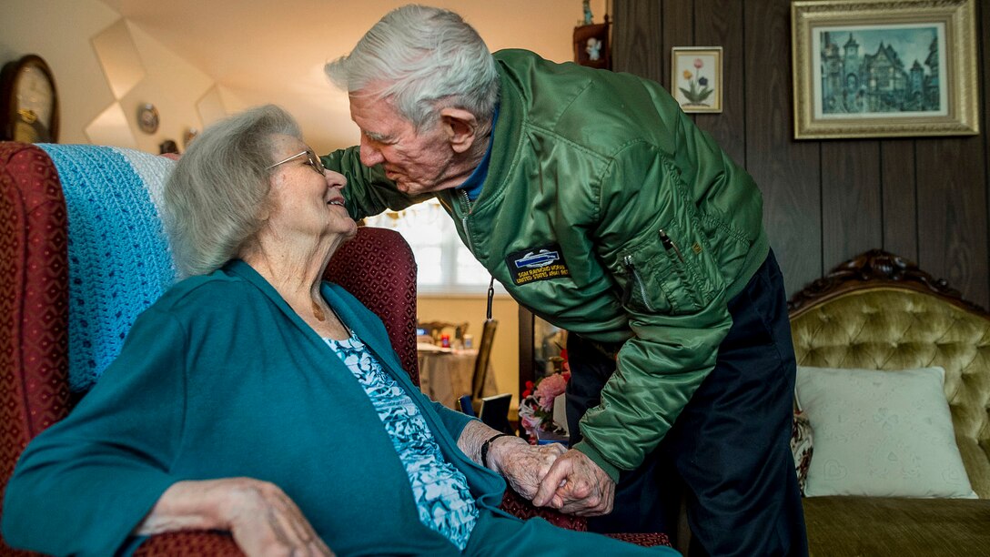 An elderly man leans in to kiss his wife, who holds her face up to his while sitting in a chair and holding his hand.