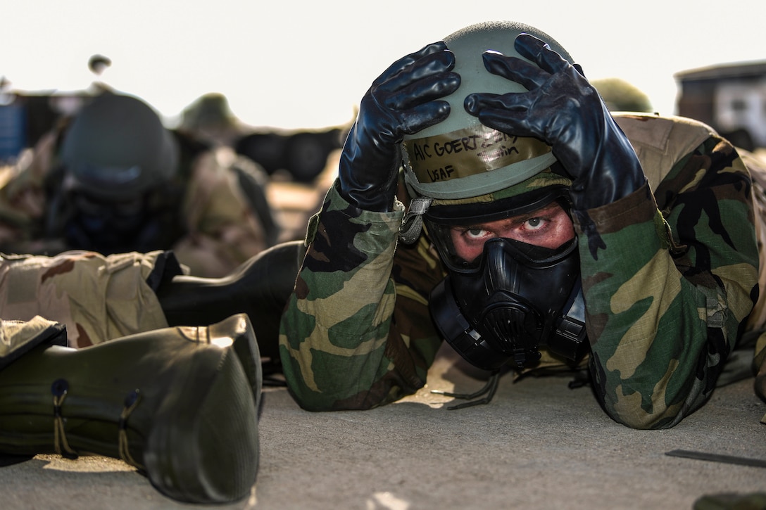 An airman in wearing a gas mask lies on the ground and places his hands on a helmet he's wearing.