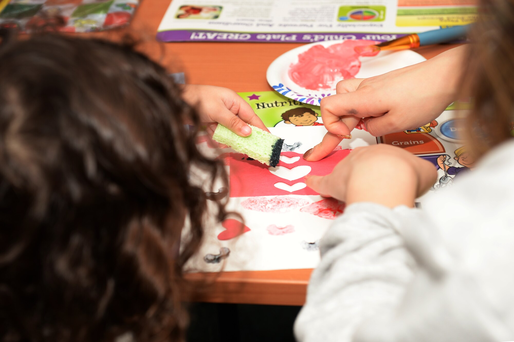 A mother helps her child sponge paint on heart-shaped paper at the Fitness and Sports Center Feb. 14, 2018 at Royal Air Force Lakenheath, England. The pair made crafts at the Veggie Valentines hosted by the Women, Infants and Children Overseas program. (U.S. Air Force photo/Airman 1st Class Shanice Williams-Jones)
