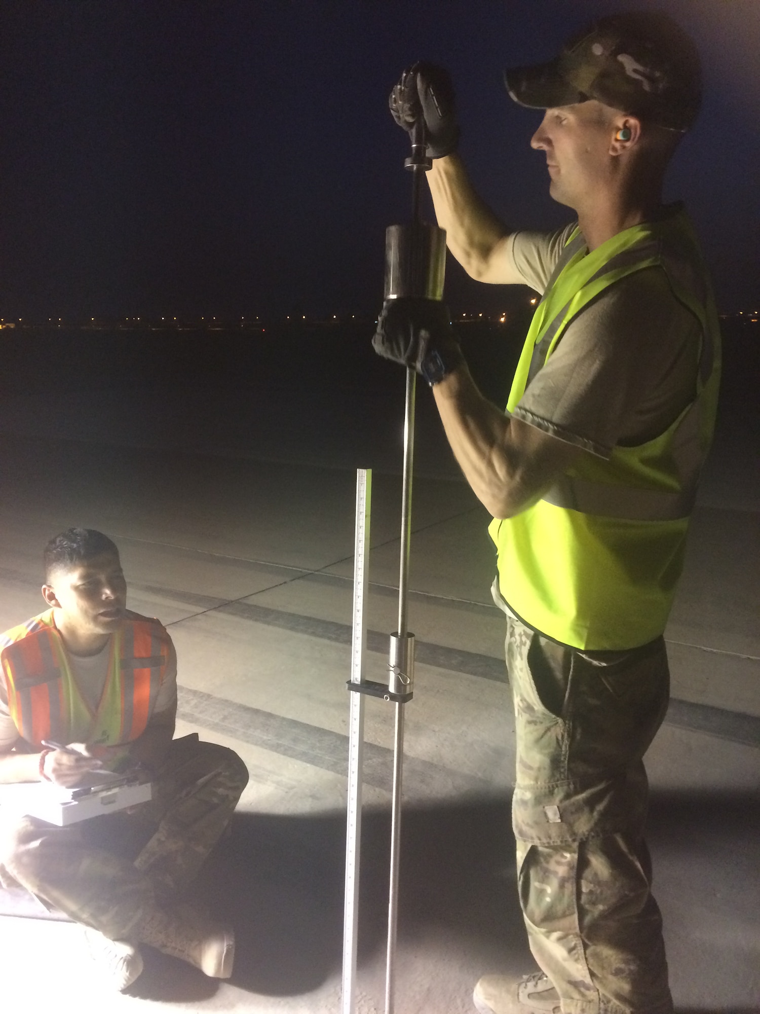 Capt Miles Ryan (standing) and TSgt Ruben Hernandez (sitting) are performing Dynamic Cone Penetrometer tests on a runway at Baghdad International Airport.  The tests were done at night to avoid high summer temperatures.