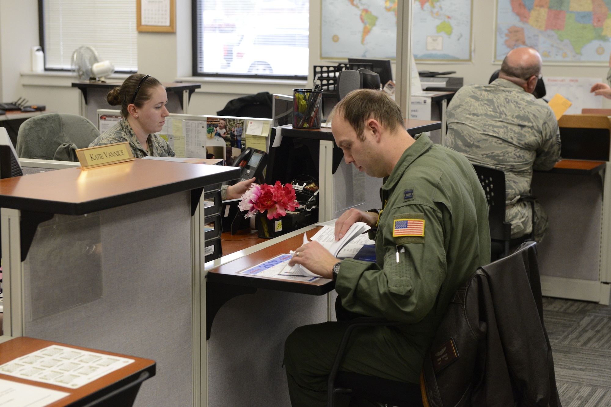 One female airman sits at a desk across from a male airman in a flight suit looking through a folder of paperwork.